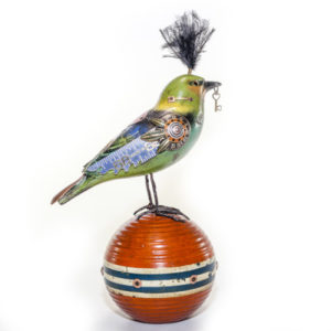 Bird with a green body and blue wings, embellished with found objects, on a red bocce ball. Created by Jim and Tori Mullan.