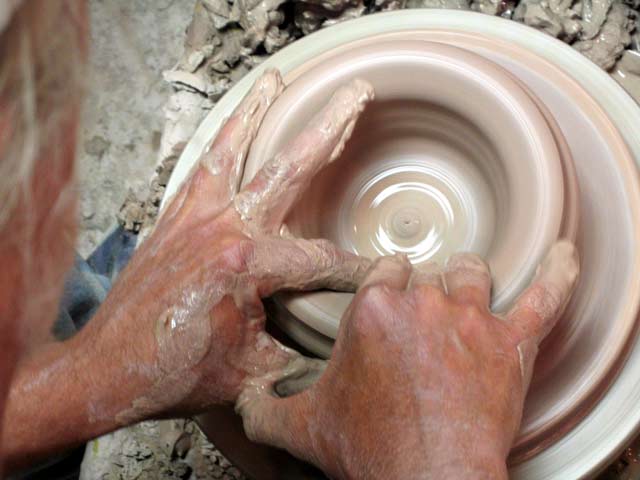Overhead closeup of clay-covered hands shaping a clay vessel on the potter's wheel