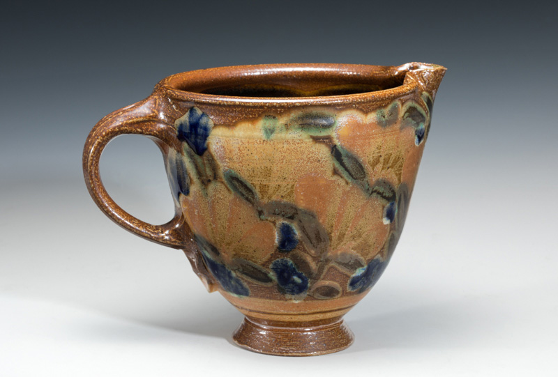 Tan ceramic pitcher with a small base and a wide body and mouth. Flowers painted onto the sides create green and blue accents.