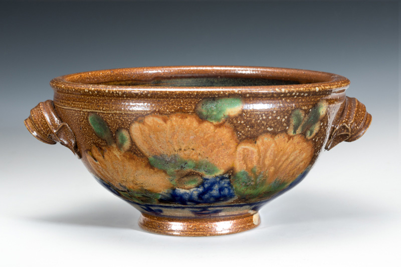 Large brown ceramic bowl with handles. Flowers painted into the glaze around the base of the bowl. Created by Terry Plasket.