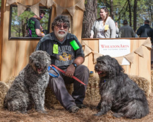 Two large leashed gray dogs sit with a person sitting on a straw bale. At PAWS for Art at WheatonArts.