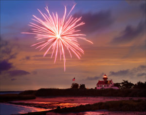 Photograph taken by Dennis Abriola of a bright pink firework exploding over the beach at sunset, with an american flag and lighthouse in the distance.