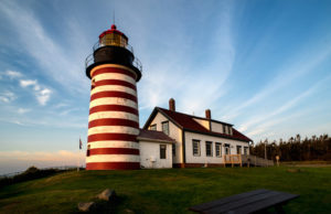 Wispy white clouds in a blue sky create a contrast against the warm colors of the large red and white striped lighthouse with a home attached to it. Photograph taken by James Evangelista