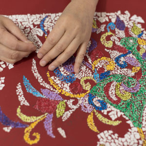 Closeup of hands assembling an abstract eggshell mosaic with white and painted shells on a red background.