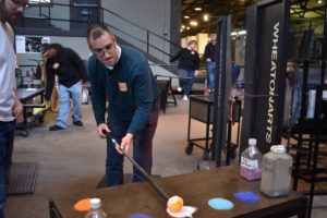 Man learning how to blow glass rolls hot glass over white powder neighboring similar powders in shades of blue.