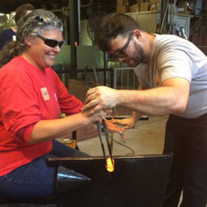 Glass Instructor, Skitch Manion, teaching a woman how to shape hot glass.