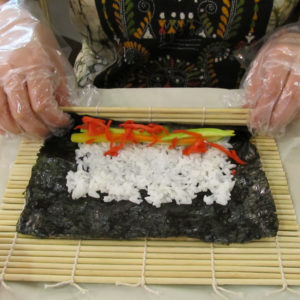 Closeup of Keiko Warner's gloved hands using a bamboo mat to roll sushi.