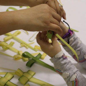 Closeup of hands guiding a child to create Philippine Palaspas with several already made palm crosses on a table