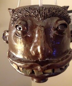 A closeup of a deep brown sculpted clay human face with jagged teeth, squinted eyes, and two small horns, created by Phyllis Seidner