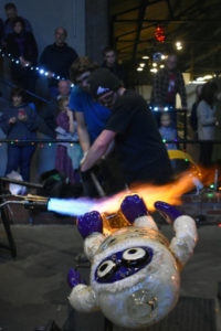 Two Big Glass Blast artists shaping a snow monster with a blowtorch. Crowd in back standing and sitting, watching intently.