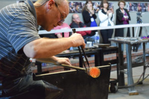 David Graeber shaping a paperweight at the end of a blowpipe, with a crowd watching behind him.