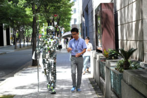 Video still of a man covered in small hexagonal mirrors walking next to a man on a city sidewalk. Created by Stanislav Müller.