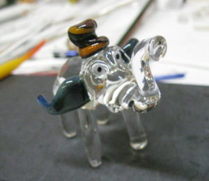 Transparent glass elephant with dark green and blue ears and a brown top hat, created by Bill Futer