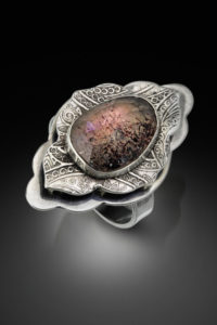 Quatrefoil shaped engraved silver ring holding a large brown and crimson gemstone. Created by Janine Krantz