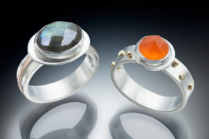 One silver right with a green gemstone and one silver ring with gold embellishments and an orange gemstone by Joan Prato