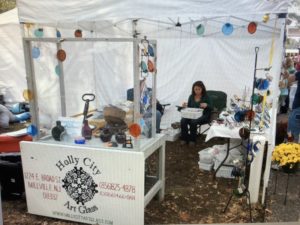 Two people sitting in James Sharpless' Holly City Art Glass booth, surrounded by a variety of items for sale.