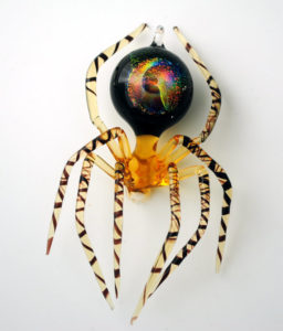 Amber colored spider with long striped legs and a multicolored, iridescent abdomen. Created by artist Thomas Von Koch