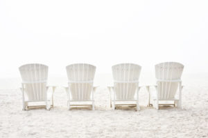 Photograph taken by Barry Hollritt of four white Adirondack chairs on a light tan beach.