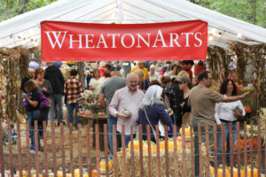 Customers explore glass pumpkin patch in a white tent, surrounded by cornstalks and straw, with a WheatonArts banner overhead