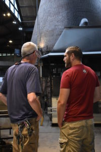 Emanation 2019 Artist Jesse Krimes speaking with Hank Adams in the Glass Studio, backs facing the camera.