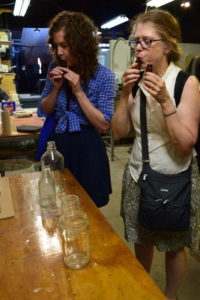 "Emanation 2019" Artists Laura Baird and Martha McDonald blow into small brown glass bottles.