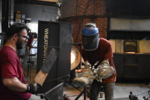 Two WheatonArts Glass Artists attach small, clear glass limbs to a larger clear glass bottle.