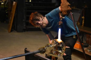 WheatonArts Glass Artist Brian R. leaning forward, using a hot torch to shape a large glass form at the end of a blowpipe