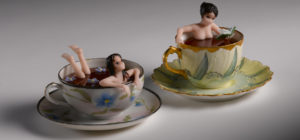 Two sculpted glass women relax inside of teacups. Art: Teacups Unfurl and Unwind, 2016, found teacup and glass, by Carmen Lozar.