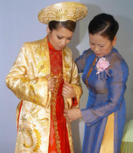 A woman in a blue gown assists a Vietnamese bride with her golden khăn đóng, ornate red gown, and golden cloak.