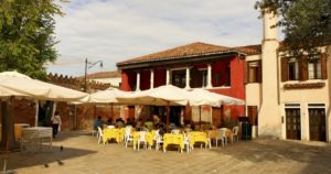 Outdoors, near the red facade of Busa Alla Torre Da Lele in Venice, people sit at yellow draped tables under white patio umbrellas