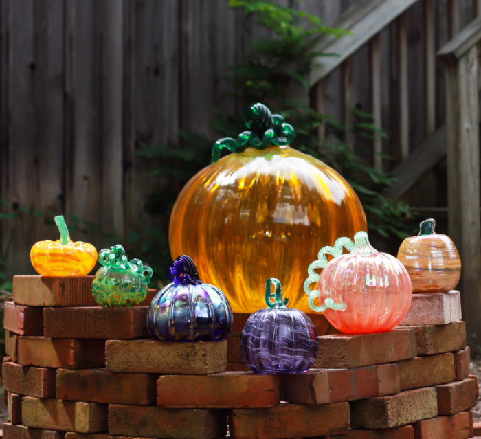 five small glass pumpkins of various colors sit on bricks in front of a large orange glass pumpkin