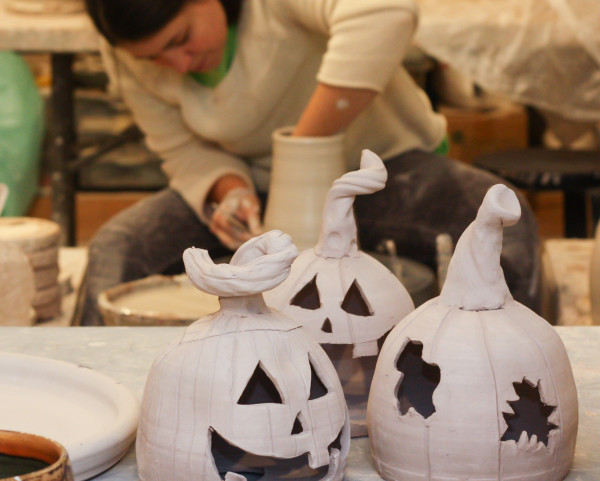 Three clay sculpted jackolanterns in the foreground with Ceramic Artist Tessa in the background forming a clay vessel on the potters wheel