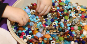 Little hands pick up a few beads from a large bowl full of glossy beads in every color, shape, and size