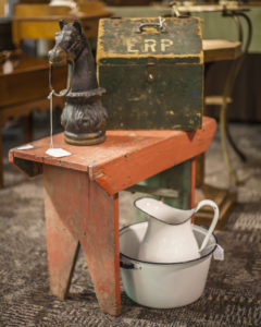 Metal horse head and a green metal box sitting on an orange wooden bench. Beneath the bench is a white pitcher and basin.