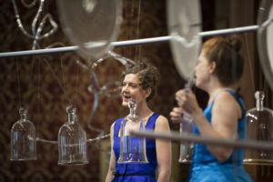 Martha McDonald and Laura Baird sing together, surrounded by suspended glass cloches and rondels for "Phantom Frequencies" at the Museum of American Glass