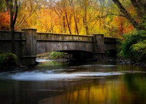 Photograph by Christopher Costa of a bridge over rippling water during autumn, with many orange and yellow trees in the background