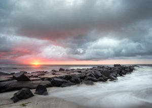 Photograph by Christopher Costa of a jetty under an orange and pink sunrise, the sky is filled with billowy clouds and fog runs over the sand.