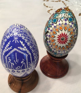 Two pysanky by Marino Diaz. The left egg has a blue nativity scene with pattern embellishments surrounding the scene. The right is decorated with a red flower and concentric multicolor shapes.