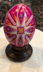 A fuchsia pysanky by Marino Diaz. The egg is decorated with a cross in the center forming a rose symbol. Images of grain and blue flowers decorate the space around it.