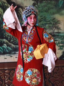 Chinese Peking Opera performer poses in a bright red outfit, carrying a box wrapped in a yellow cloth.