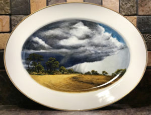 A painted porcelain plate with a scene of a storm cloud looming over the plains. Created by Michelle Post.