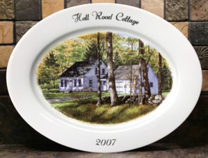 A painted porcelain plate with a scene of a home with a barn beside it, slightly obscured by trees in the large forest around it. Text at the top reads Hall Road Cottage, bottom text reading 2007. Created by Michelle Post