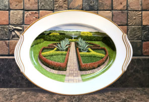 A painted porcelain plate with a scene of a well manicured garden with tall hedges and leafy green plants, a peek of a lake in the distance. Created by Michelle Post