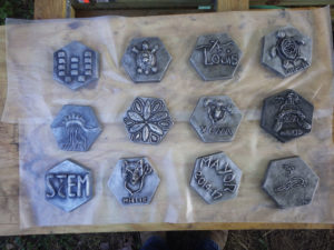 12 metal hexagons with various designs laid onto a wooden table. Created by Rachel Marie Wenner.