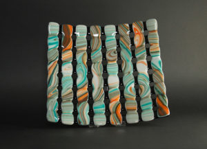 Glass piece by Stephanie Baness made up of 8 columns colored in a marbled pattern of orange, teal, and white, connected with 8 rows of black behind.