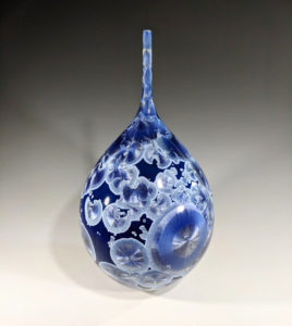 Blue ceramic piece by Erika Novak and Drew Darley with a wide body tapering to a long cylinder, cell-like silver and blue circles throughout the piece