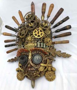 A found object mask by Larry Agnello, referencing masks from Africa. Its headdress is made up of gears and a variety of tools with wooden handles sticking out like feathers.