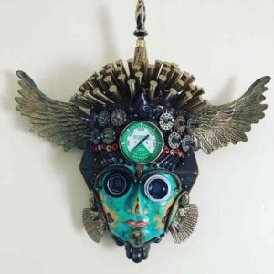 A found object mask by Larry Agnello. Wings on either side of the head, an oxygen meter in the center of the forehead, and a full green face with camera lenses for eyes.