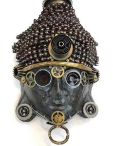 A found object mask by Larry Agnello, referencing masks from Africa. Metal gears and rings adorn its deep green face,