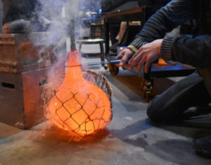 A large gather of molten glass is blown into a chain link cage